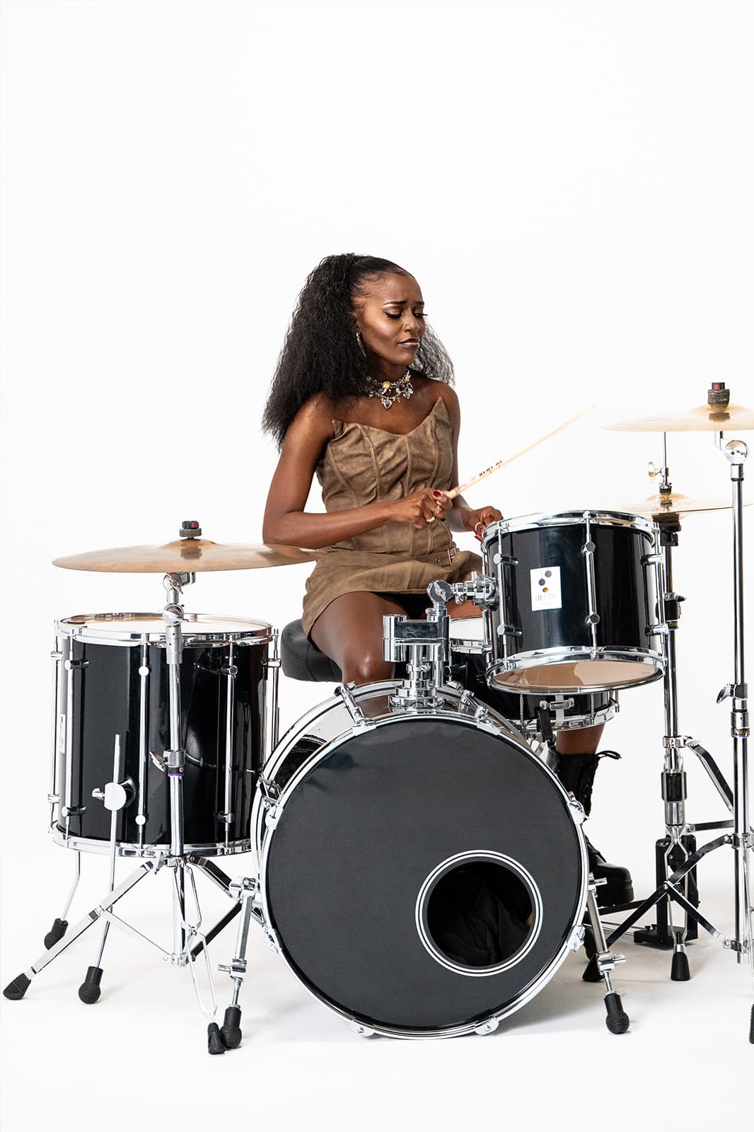 Becky Sangolo playing a black drum kit in a tan leather dress during a studio shoot