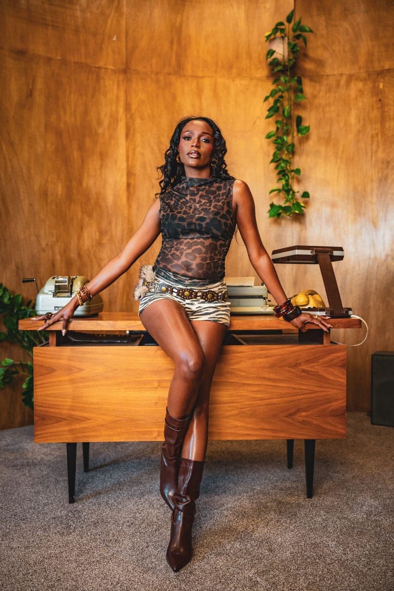Becky Sangolo in a leopard-print top and fur-trimmed mini skirt sitting on a mid-century wooden desk with a vintage typewriter and telephone