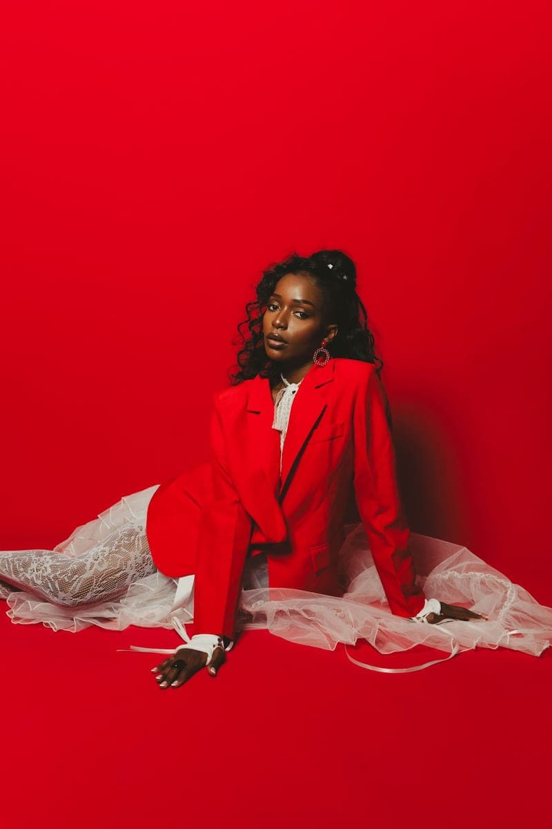 Becky Sangolo seated on the floor in a red blazer and white tulle skirt against a red backdrop